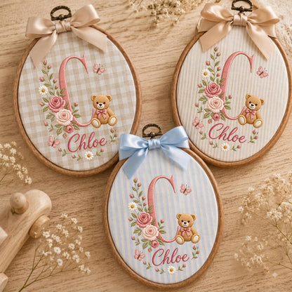 May include: Three oval wooden embroidery hoops featuring fabric backgrounds with a large embroidered letter C, a teddy bear, pink roses, daisies, and butterflies. Each hoop is topped with a satin ribbon bow and displays the name Chloe in script.