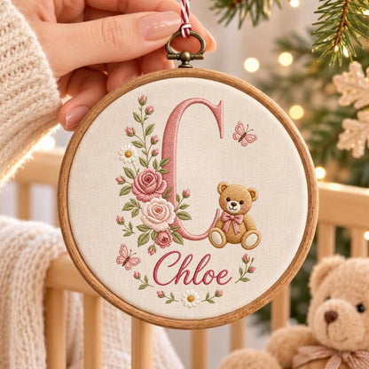 May include: A circular wooden embroidery hoop featuring a cream-colored fabric background. The design includes a large pink embroidered letter C, a brown teddy bear with a pink bow, pink roses, green leaves, white daisies, and two butterflies.