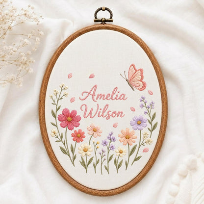 May include: An oval wooden embroidery hoop containing white fabric with hand-stitched floral designs in pink, purple, and yellow. The text Amelia Wilson is embroidered in pink script in the center, accompanied by a pink butterfly and scattered petals.