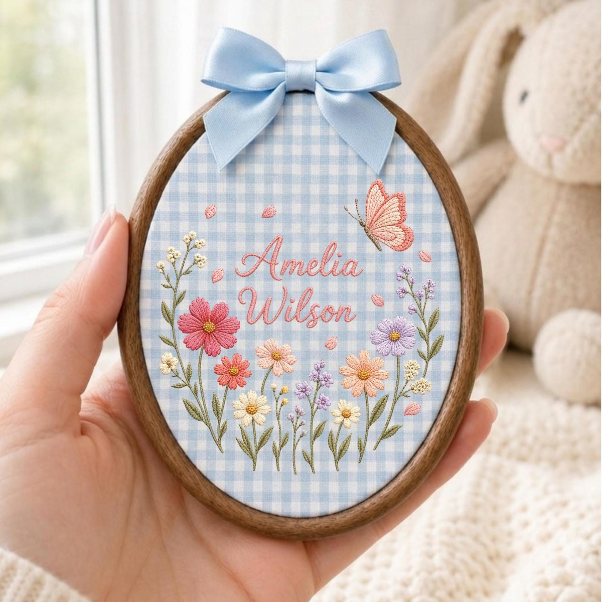 May include: An oval embroidery hoop with a dark wood frame containing light blue gingham fabric. The fabric features embroidered flowers in pink, orange, and purple, a small butterfly, and the text Amelia Wilson in pink cursive script.