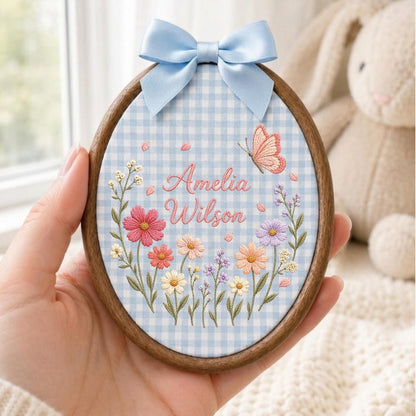 May include: An oval embroidery hoop with a dark wood frame containing light blue gingham fabric. The fabric features embroidered flowers in pink, orange, and purple, a small butterfly, and the text Amelia Wilson in pink cursive script.