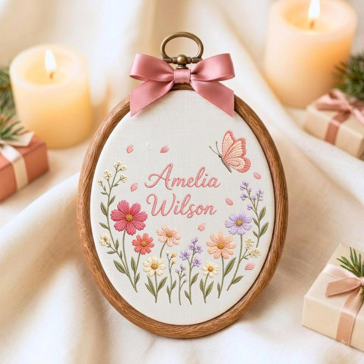 May include: An oval wooden embroidery hoop containing white fabric with the embroidered name Amelia Wilson in pink script. The design features colorful wildflowers in shades of pink, purple, and orange, a pink butterfly, and a pink satin bow.