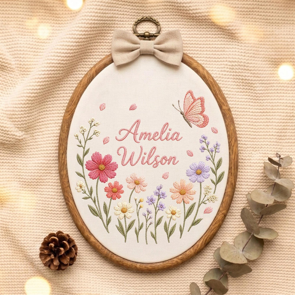May include: An oval embroidery hoop featuring a white fabric center with the embroidered name Amelia Wilson in pink script. The design includes colorful floral stems in shades of pink, purple, and yellow, a pink butterfly, and a beige fabric bow.