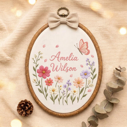 May include: An oval embroidery hoop featuring a white fabric center with the embroidered name Amelia Wilson in pink script. The design includes colorful floral stems in shades of pink, purple, and yellow, a pink butterfly, and a beige fabric bow.