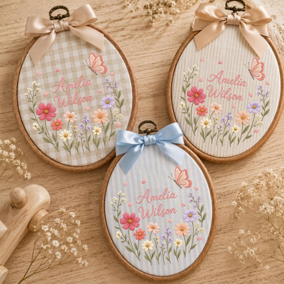May include: Three oval wooden embroidery hoops featuring fabric backgrounds with gingham or striped patterns. Each hoop displays embroidered floral arrangements, a butterfly, and the name Amelia Wilson in pink script, topped with a satin bow.