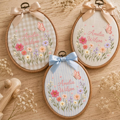 May include: Three oval wooden embroidery hoops featuring fabric backgrounds with gingham or striped patterns. Each hoop displays embroidered floral arrangements, a butterfly, and the name Amelia Wilson in pink script, topped with a satin bow.