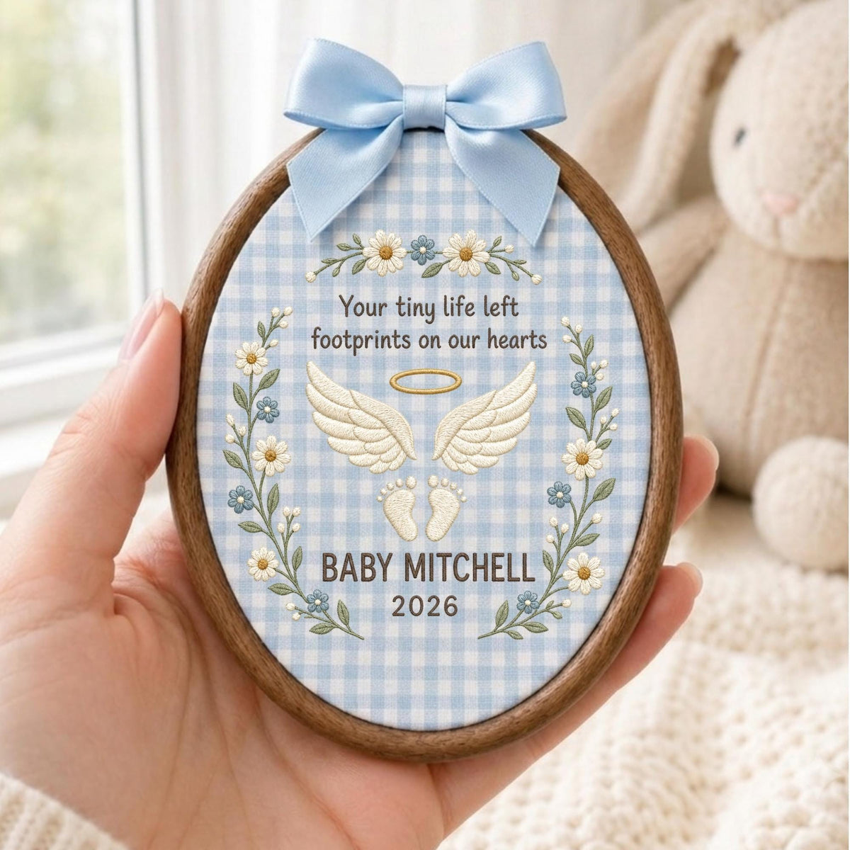 May include: An oval wooden embroidery hoop containing light blue and white gingham fabric. The fabric features embroidered white daisy vines, a golden halo, white angel wings, baby footprints, and the text: Your tiny life left footprints on our hearts, BABY MITCHELL, 2026. A light blue satin bow is attached to the top.