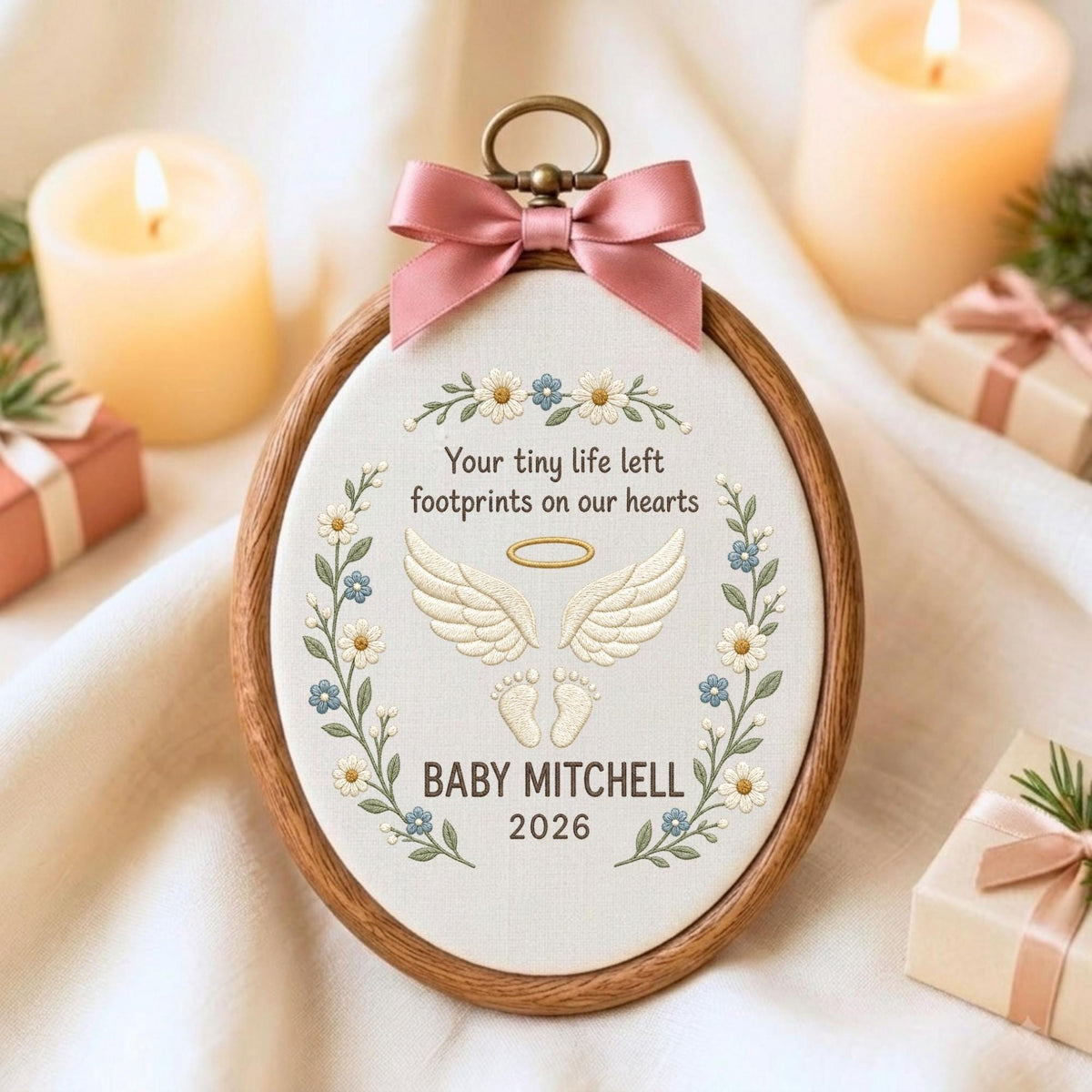May include: An oval wooden embroidery hoop containing white fabric with floral embroidery, a pair of angel wings, a halo, and baby footprints. The text reads: Your tiny life left footprints on our hearts, BABY MITCHELL, 2026. A pink satin bow is attached to the top.