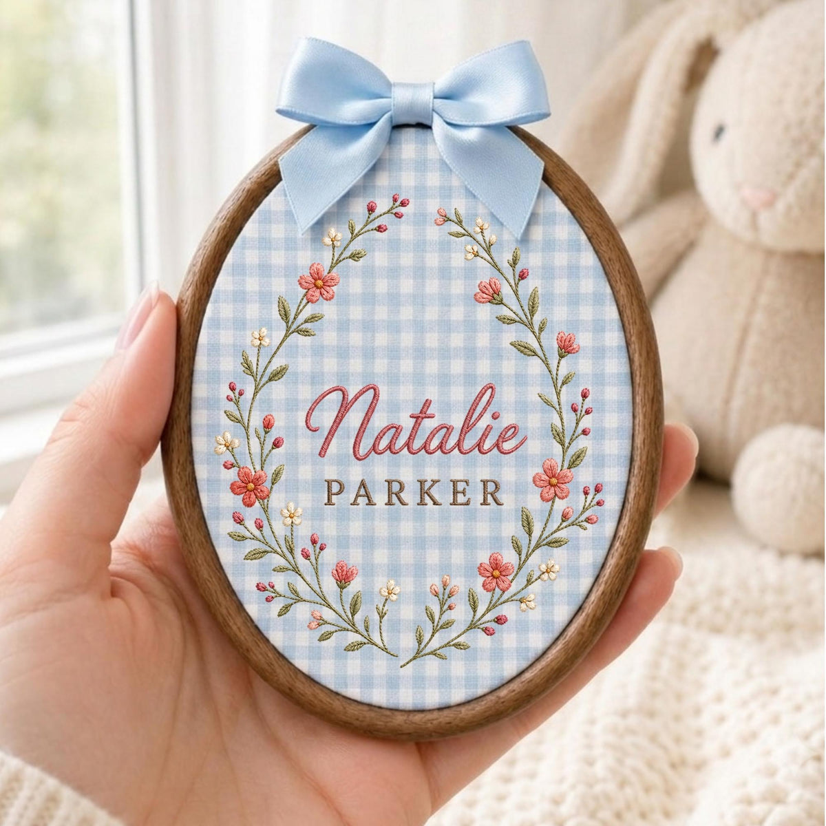 May include: An oval wooden embroidery hoop containing light blue and white gingham fabric. The fabric features an embroidered floral wreath in pink and green, surrounding the name Natalie Parker in pink and brown text, topped with a light blue satin bow.