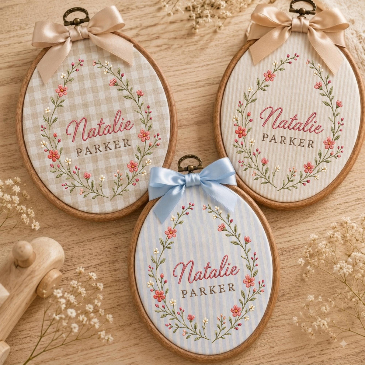 May include: Three oval wooden embroidery hoops featuring fabric backgrounds in beige gingham, white stripes, and light blue stripes. Each hoop displays an embroidered floral wreath surrounding the name Natalie Parker and is topped with a satin bow.