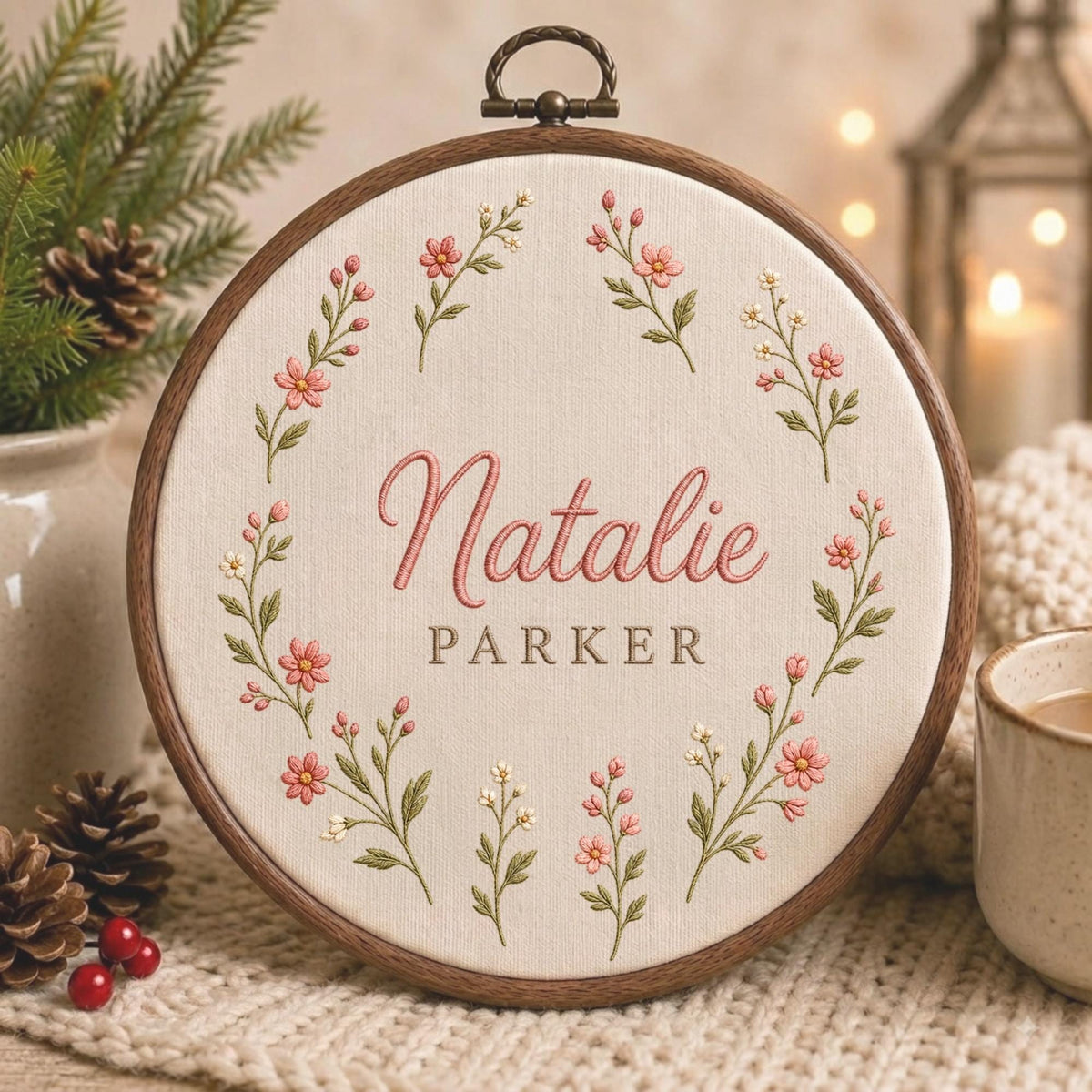 May include: A circular wooden embroidery hoop containing cream-colored fabric with a floral wreath design. The embroidery features pink and white flowers with green stems. The name Natalie is stitched in pink script, with the name PARKER in brown.