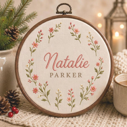 May include: A circular wooden embroidery hoop containing cream-colored fabric with a floral wreath design. The embroidery features pink and white flowers with green stems. The name Natalie is stitched in pink script, with the name PARKER in brown.