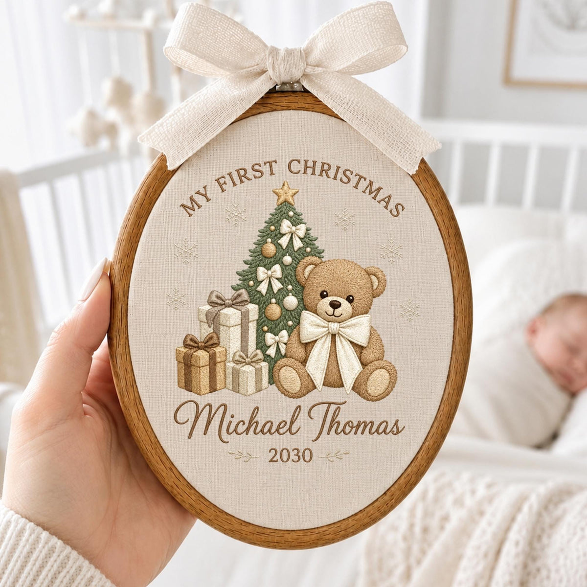 May include: An oval wooden embroidery hoop containing a beige fabric panel with a printed design of a teddy bear, a Christmas tree, and gift boxes. The text reads "MY FIRST CHRISTMAS", "Michael Thomas", and "2030". A beige ribbon bow is attached.