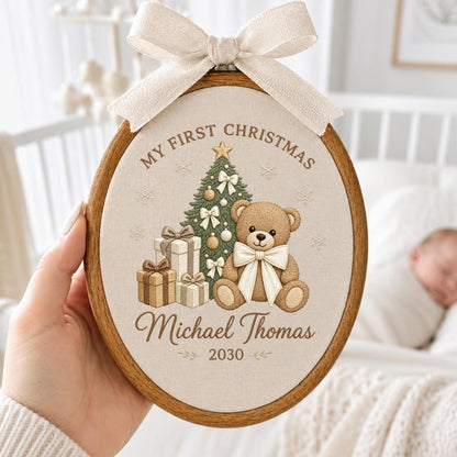 May include: An oval wooden embroidery hoop containing a beige fabric panel with a printed design of a teddy bear, a Christmas tree, and gift boxes. The text reads "MY FIRST CHRISTMAS", "Michael Thomas", and "2030". A beige ribbon bow is attached.
