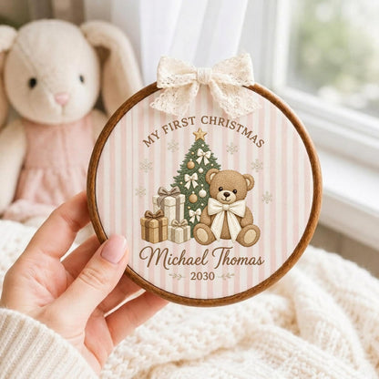 May include: A round wooden embroidery hoop frame containing fabric with a light pink and white striped pattern. The design features a teddy bear, a Christmas tree, gift boxes, and the text "MY FIRST CHRISTMAS Michael Thomas 2030". A cream lace bow is attached to the top.