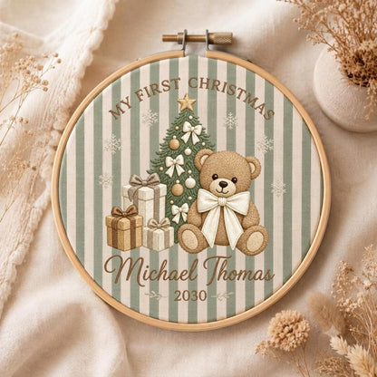 May include: A round wooden embroidery hoop framing fabric with sage green and white vertical stripes. The design features a brown teddy bear, a decorated Christmas tree, and gift boxes. Text reads: MY FIRST CHRISTMAS, Michael Thomas, 2030.