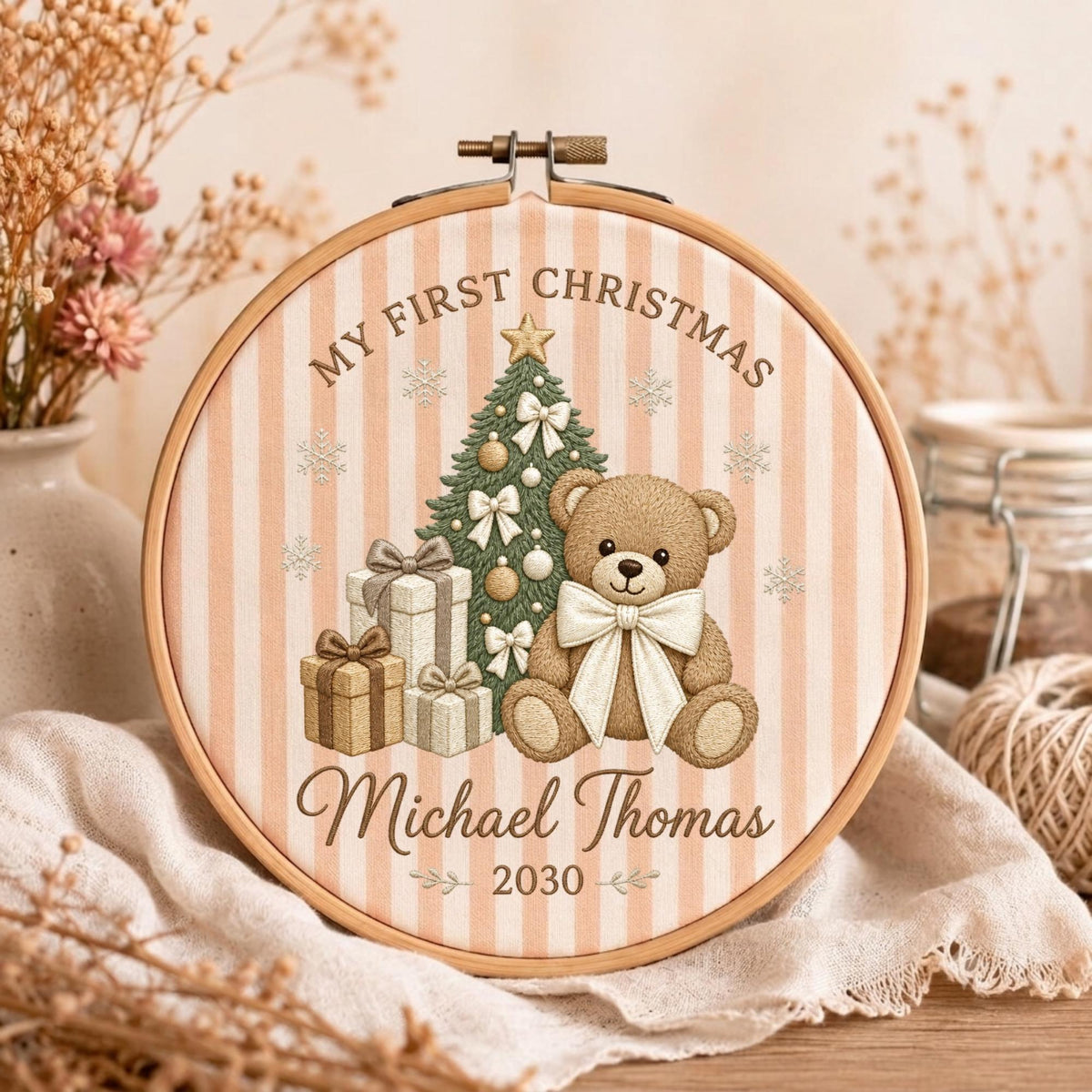 May include: A circular wooden embroidery hoop framing fabric with vertical beige stripes. The design features a teddy bear, a decorated Christmas tree, and gift boxes. Text reads: MY FIRST CHRISTMAS, Michael Thomas, and the year 2030.