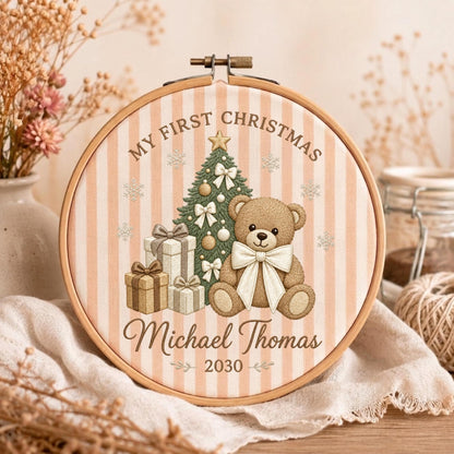 May include: A circular wooden embroidery hoop framing fabric with vertical beige stripes. The design features a teddy bear, a decorated Christmas tree, and gift boxes. Text reads: MY FIRST CHRISTMAS, Michael Thomas, and the year 2030.
