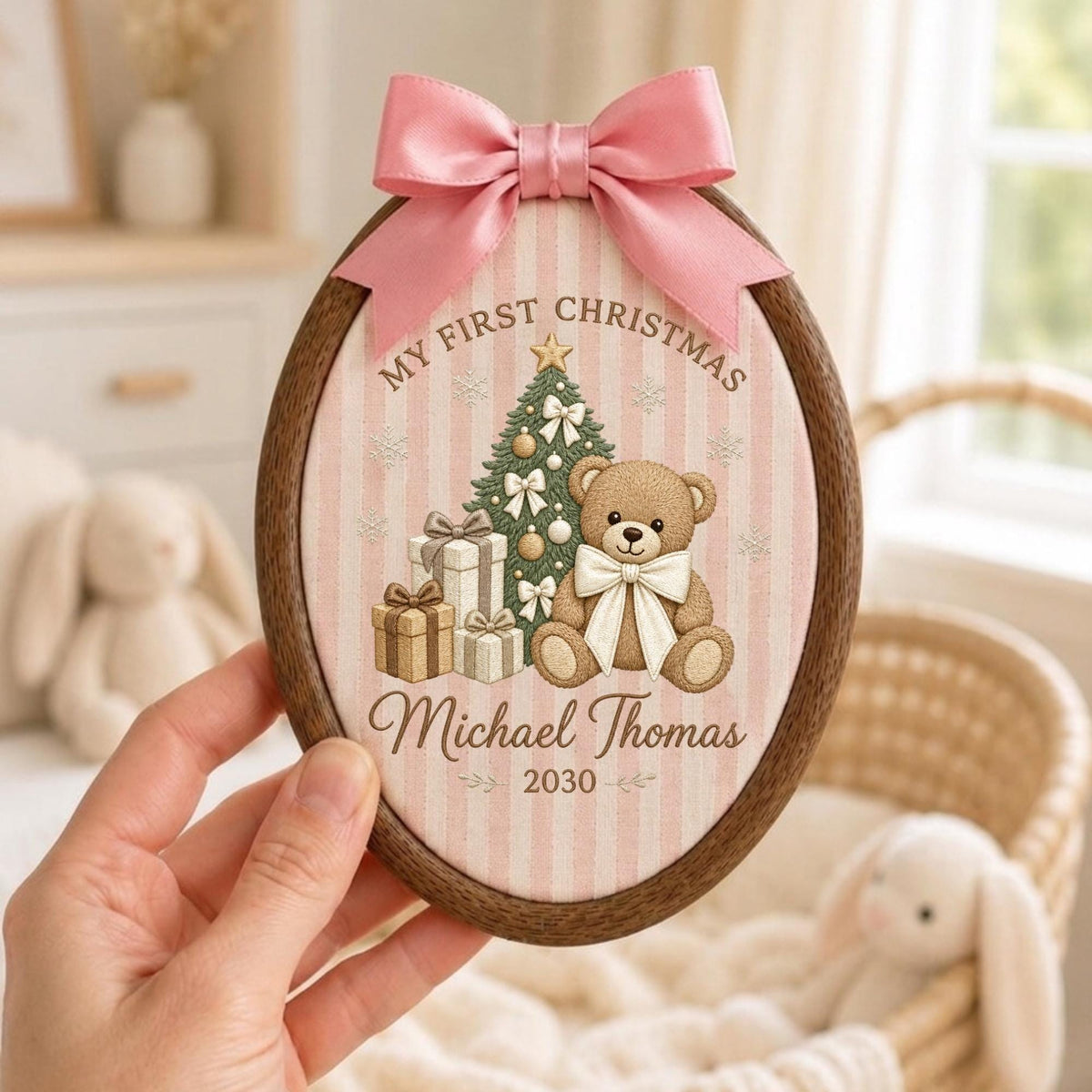 May include: An oval wooden embroidery hoop frame containing fabric with a pink and white striped pattern. The design features a teddy bear, a Christmas tree, and gifts with the text "MY FIRST CHRISTMAS", "Michael Thomas", and "2030". A pink satin bow is attached to the top.