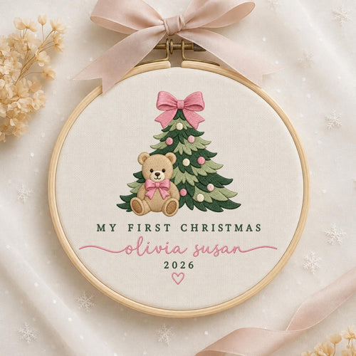 May include: A round wooden embroidery hoop featuring a cream fabric background with a stitched green Christmas tree, a light brown teddy bear with a pink bow, and the text MY FIRST CHRISTMAS, olivia susan, 2026, and a small pink heart.