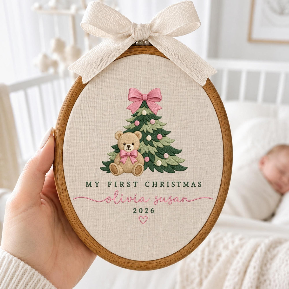 May include: An oval wooden embroidery hoop containing cream-colored fabric with an embroidered green Christmas tree, a tan teddy bear with a pink bow, and the text "MY FIRST CHRISTMAS", "olivia susan", "2026", and a small heart, topped with a beige ribbon.