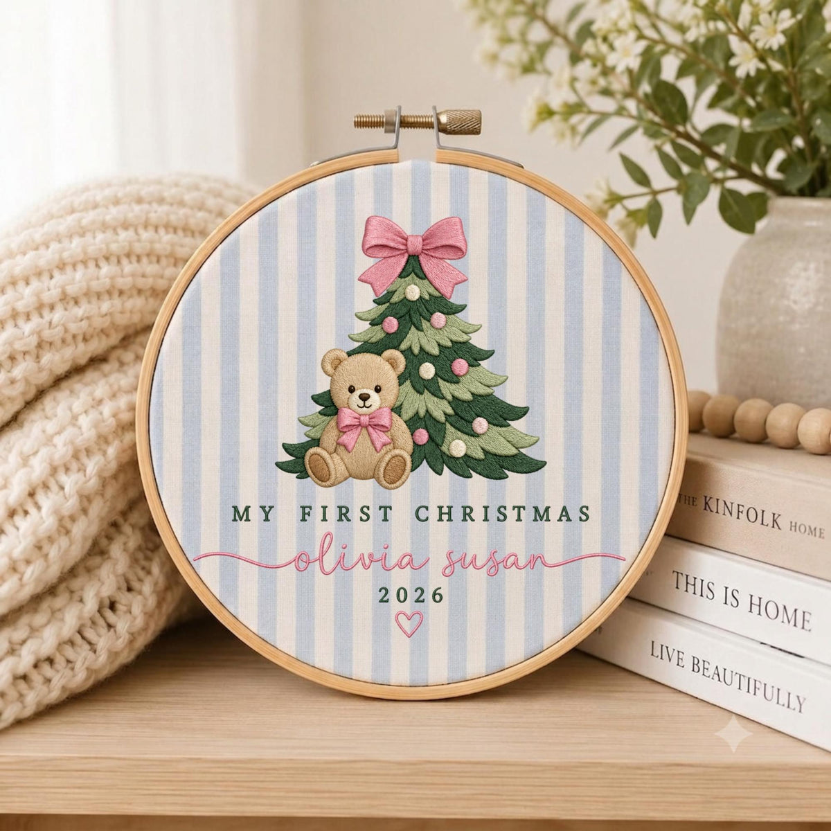 May include: A circular embroidery hoop featuring a light blue and white striped fabric background. The design includes a green Christmas tree with pink ornaments, a pink bow, and a tan teddy bear wearing a pink bow. Text reads MY FIRST CHRISTMAS, Olivia Susan, 2026, and a small pink heart.