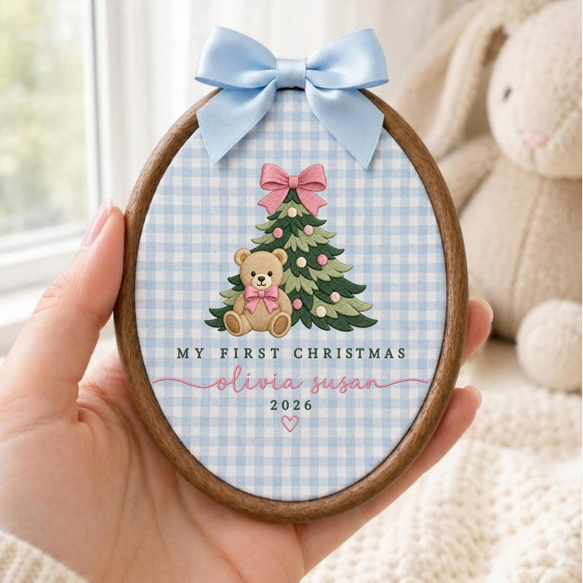 May include: An oval wooden embroidery hoop ornament featuring a light blue gingham fabric background. It displays an embroidered Christmas tree with a pink bow, a teddy bear with a pink bow, and the text My First Christmas Olivia Susan 2026.