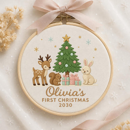 May include: A round wooden embroidery hoop containing fabric with a stitched design of a green Christmas tree, a brown deer, a squirrel, and a rabbit. Below are three gift boxes and the text Olivia's FIRST CHRISTMAS 2030. A light pink ribbon is tied at the top.