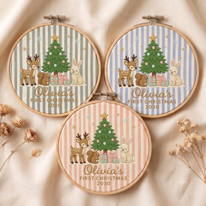 May include: Three circular embroidery hoops featuring fabric with vertical stripes in sage green, light blue, and soft peach. Each hoop displays an embroidered Christmas tree, a deer, a squirrel, a rabbit, and gifts with the text Olivia's FIRST CHRISTMAS 2030.
