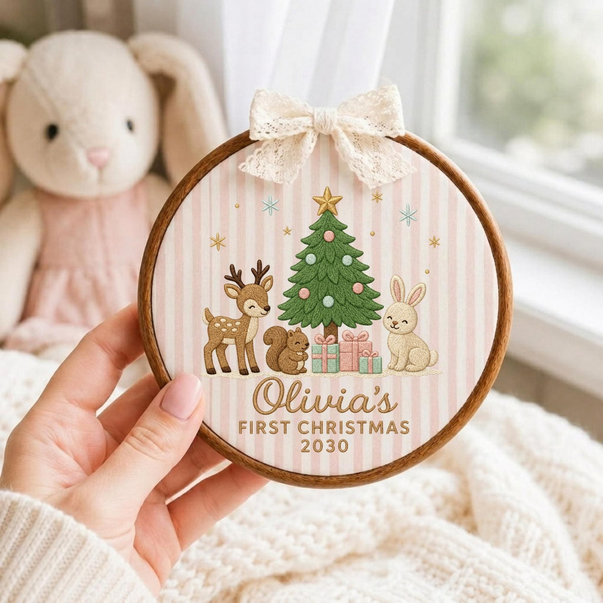 May include: A round wooden embroidery hoop frame containing fabric with a pink and white striped pattern. The design features a Christmas tree, a deer, a squirrel, and a rabbit with the text Olivia's FIRST CHRISTMAS 2030. A cream lace bow is attached to the top.