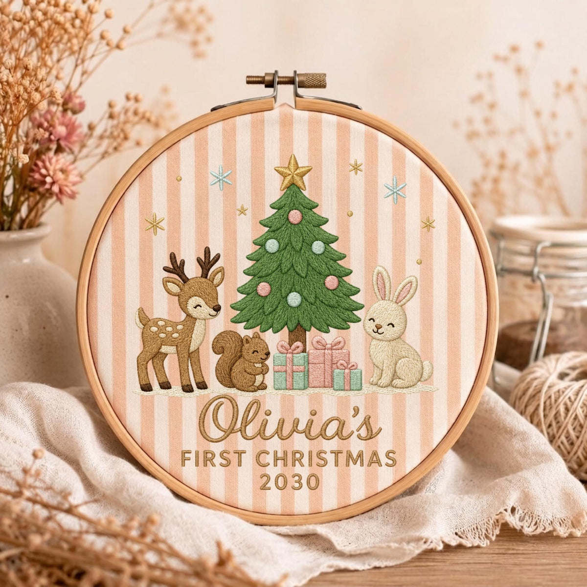 May include: A round wooden embroidery hoop containing fabric with vertical beige stripes. The embroidery features a green Christmas tree, a deer, a squirrel, a rabbit, and gifts. Text reads: Olivia's FIRST CHRISTMAS 2030, stitched in brown thread.