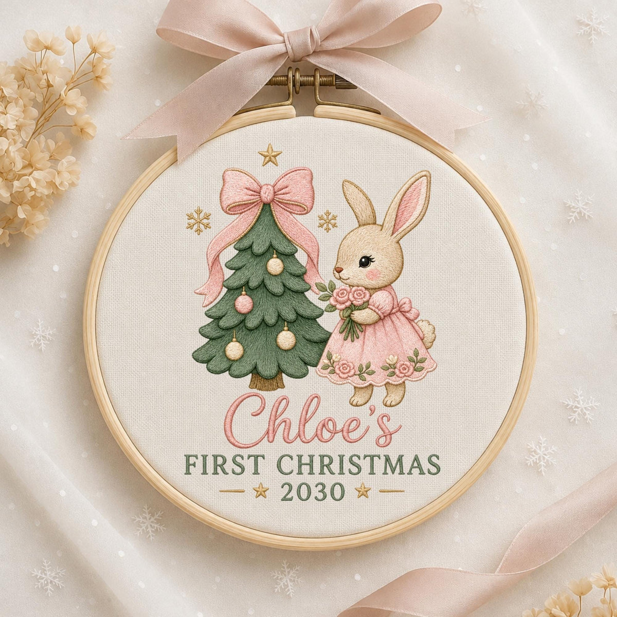 May include: A round wooden embroidery hoop containing fabric with a stitched design of a green Christmas tree and a beige rabbit in a pink dress holding flowers. The text reads Chloe's FIRST CHRISTMAS 2030. A light pink ribbon is tied at the top.
