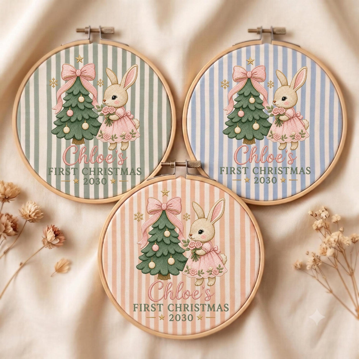 May include: Three circular wooden embroidery hoops featuring fabric with vertical stripes in sage green, light blue, and peach. Each displays a cartoon rabbit in a pink dress next to a decorated Christmas tree with the text Chloe's FIRST CHRISTMAS 2030.