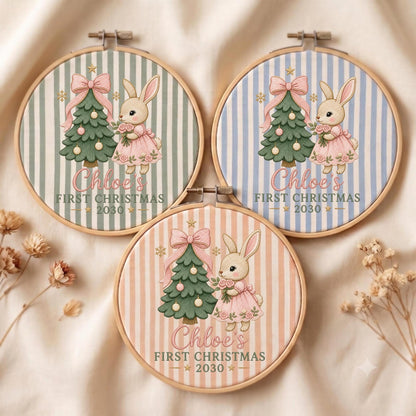 May include: Three circular wooden embroidery hoops featuring fabric with vertical stripes in sage green, light blue, and peach. Each displays a cartoon rabbit in a pink dress next to a decorated Christmas tree with the text Chloe's FIRST CHRISTMAS 2030.
