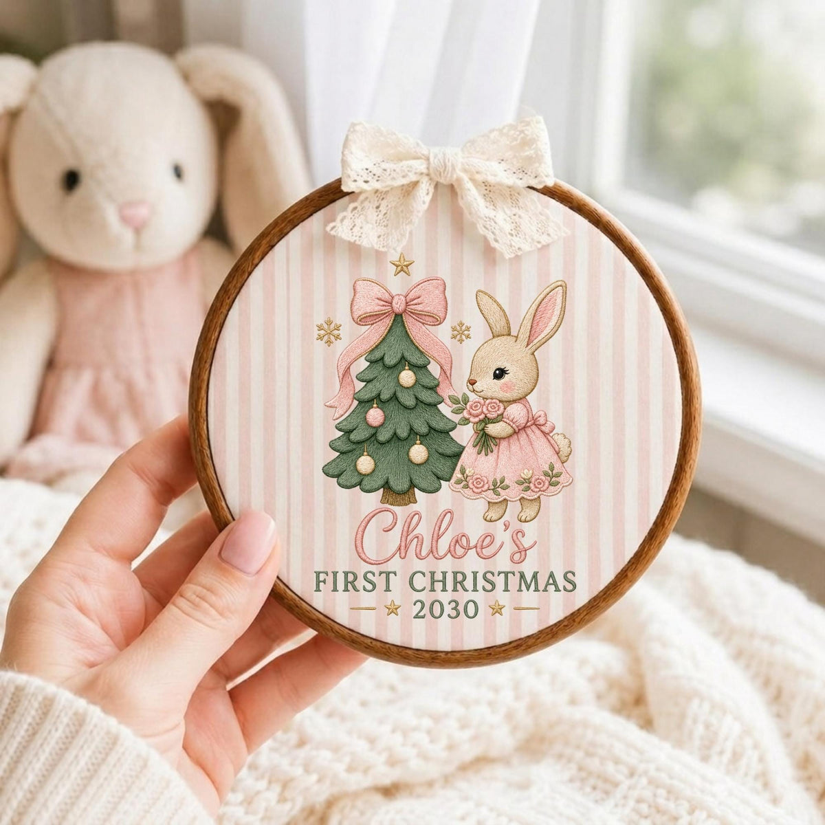 May include: A circular wooden embroidery hoop ornament featuring a light pink and white striped fabric background. The design includes a green Christmas tree, a rabbit in a pink dress, and the text Chloe's FIRST CHRISTMAS 2030. A cream lace bow is attached to the top.