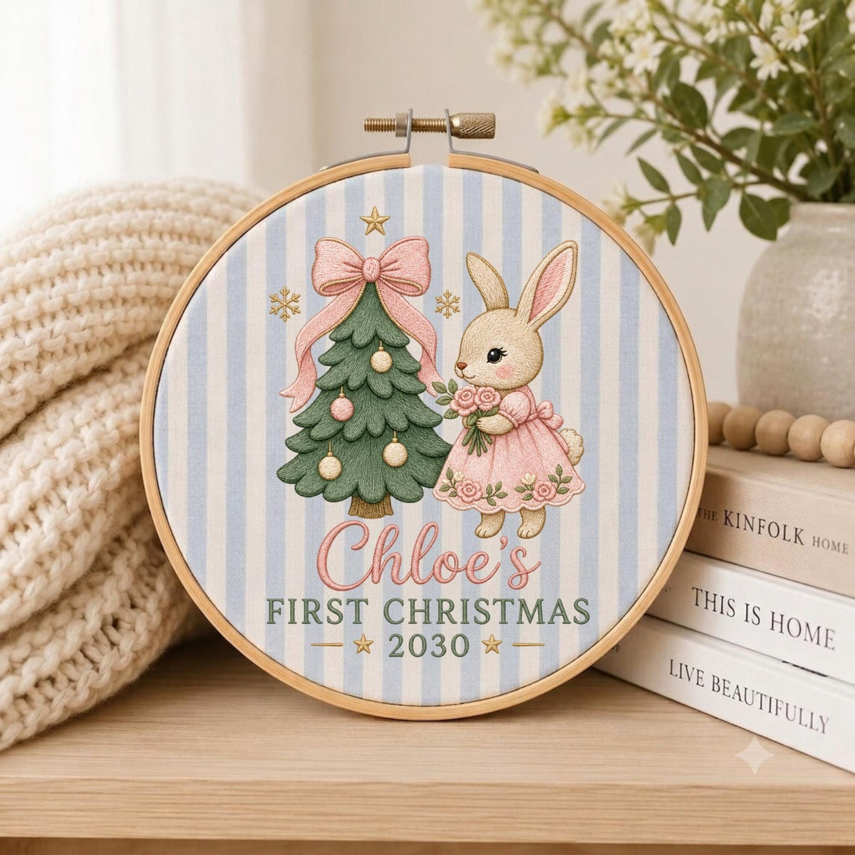May include: A round wooden embroidery hoop containing fabric with a light blue and white vertical stripe pattern. The design features a green Christmas tree with a pink bow, a rabbit in a pink dress, and the text Chloe's FIRST CHRISTMAS 2030.