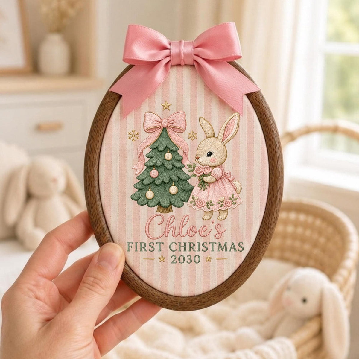 May include: An oval wooden embroidery hoop frame containing fabric with a pink and white striped pattern. The design features a green Christmas tree, a rabbit in a pink dress, and the text Chloe's FIRST CHRISTMAS 2030. A pink satin bow is attached to the top.