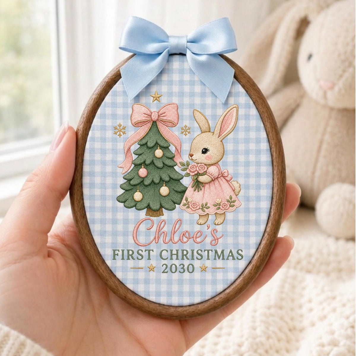 May include: An oval wooden embroidery hoop ornament featuring a light blue gingham fabric background. The design includes a green Christmas tree with pink ornaments, a rabbit in a pink dress, and the text Chloe's FIRST CHRISTMAS 2030.