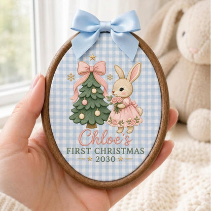 May include: An oval wooden embroidery hoop ornament featuring a light blue gingham fabric background. The design includes a green Christmas tree with pink ornaments, a rabbit in a pink dress, and the text Chloe's FIRST CHRISTMAS 2030.