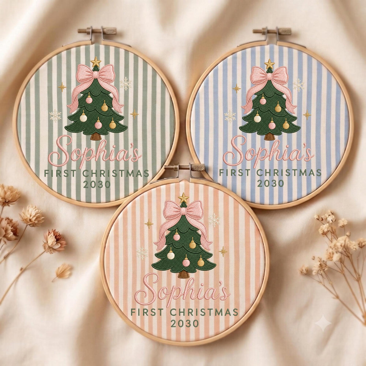 May include: Three circular wooden embroidery hoops featuring fabric with vertical stripes in sage green, light blue, and peach. Each displays a green Christmas tree with a pink bow, gold star, and ornaments, with the text Sophia's FIRST CHRISTMAS 2030.