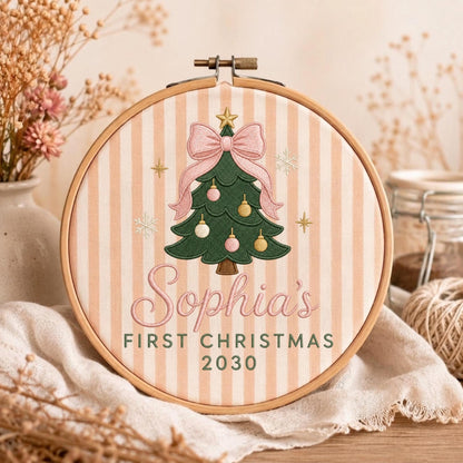 May include: A round wooden embroidery hoop containing fabric with vertical beige stripes. The design features a green Christmas tree with a large pink bow, pink and gold ornaments, and a gold star. Text reads: Sophia's FIRST CHRISTMAS 2030.