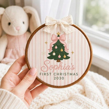 May include: A circular wooden embroidery hoop frame containing fabric with a light pink and white vertical striped pattern. The fabric features a green Christmas tree illustration with pink and gold ornaments, a pink bow, and the text Sophia's FIRST CHRISTMAS 2030. A cream-colored lace bow is attached to the top.