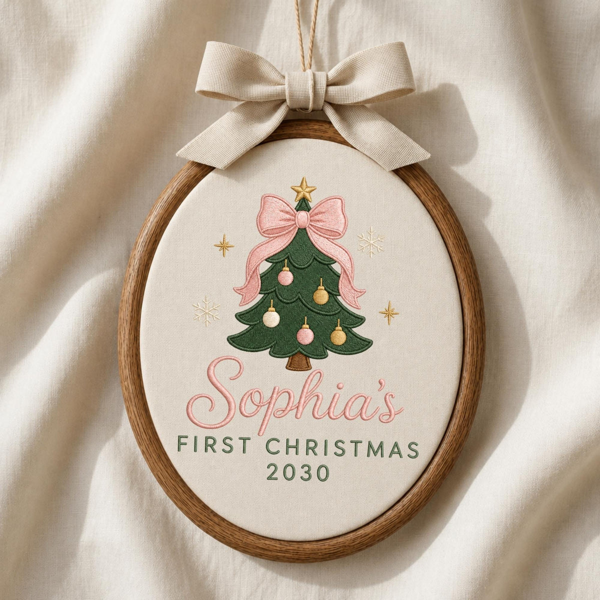 May include: An oval embroidery hoop ornament featuring a cream fabric background with a green Christmas tree illustration, pink and gold ornaments, a pink bow, and a gold star. The text reads Sophia's FIRST CHRISTMAS 2030. A beige ribbon bow is attached to the top.