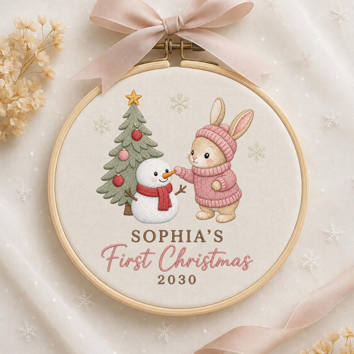 May include: A round wooden embroidery hoop containing fabric with a stitched design of a rabbit in a pink sweater and a snowman next to a Christmas tree. The text reads SOPHIA'S First Christmas 2030. A light pink ribbon is tied at the top.