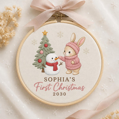 May include: A round wooden embroidery hoop containing fabric with a stitched design of a rabbit in a pink sweater and a snowman next to a Christmas tree. The text reads SOPHIA'S First Christmas 2030. A light pink ribbon is tied at the top.