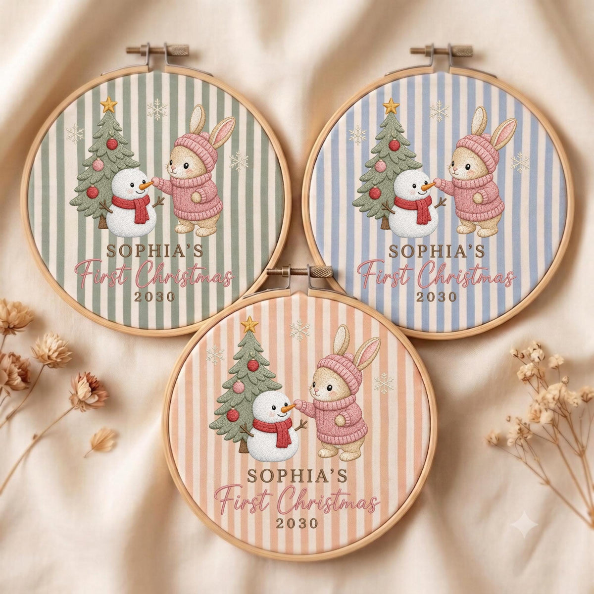 May include: Three circular wooden embroidery hoops featuring fabric with vertical stripes in sage green, light blue, and peach. Each displays a bunny in a pink sweater, a snowman, a Christmas tree, and the text Sophia's First Christmas 2030.