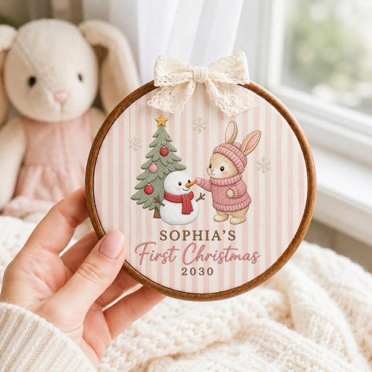 May include: A round wooden embroidery hoop ornament featuring a light pink and white striped fabric background. The design shows a bunny in a pink sweater, a snowman, and a Christmas tree. Text reads SOPHIA'S First Christmas 2030. A cream lace bow is attached to the top.