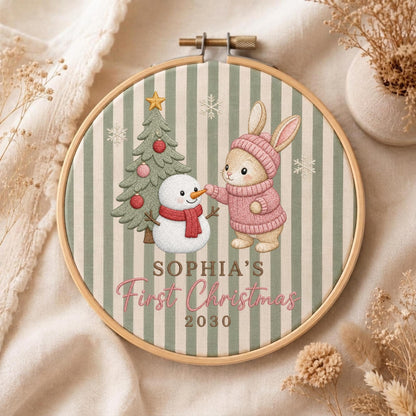 May include: A circular embroidery hoop featuring a green and white striped fabric background. The design includes a decorated Christmas tree, a snowman, and a rabbit wearing a pink sweater and hat. Text reads: SOPHIA'S First Christmas 2030.