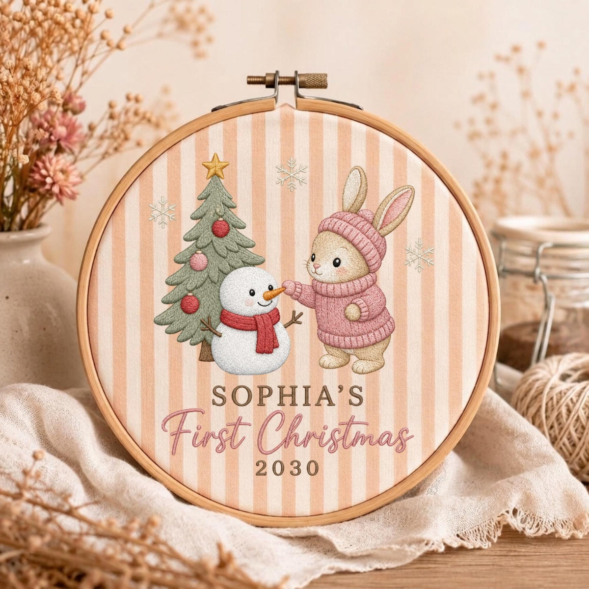 May include: A round wooden embroidery hoop containing fabric with vertical beige stripes. The design features a green Christmas tree, a white snowman with a red scarf, and a brown rabbit in a pink sweater. Text reads: SOPHIA'S First Christmas 2030.