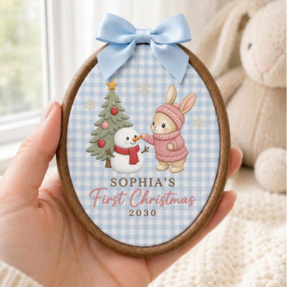 May include: An oval wooden embroidery hoop ornament featuring a light blue gingham fabric background. The design shows a bunny in a pink sweater, a snowman, and a Christmas tree. Text reads: SOPHIA'S First Christmas 2030. A light blue satin bow is attached to the top.